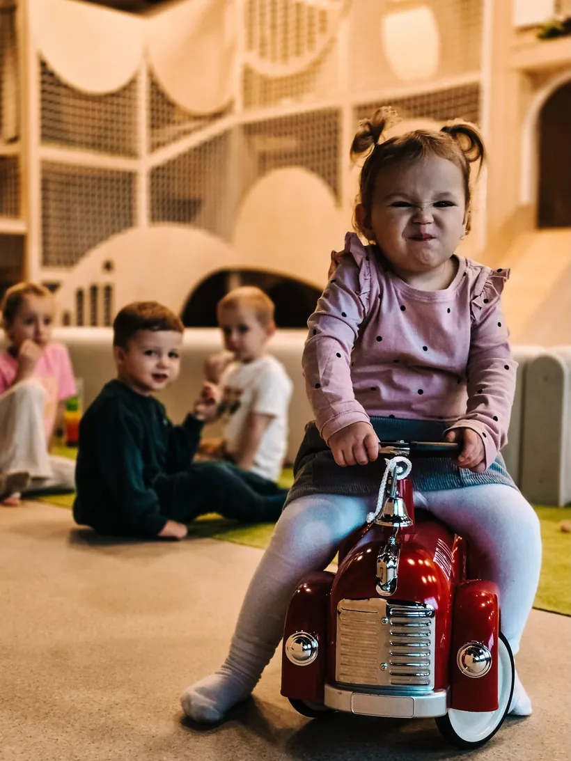 Dziewczynka na czerwonym jeździku, w tle inne dzieci
